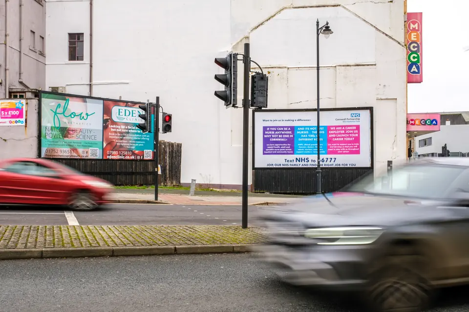 Two billboards on Fore Street, Plymouth, beside the traffic lights as cars pass